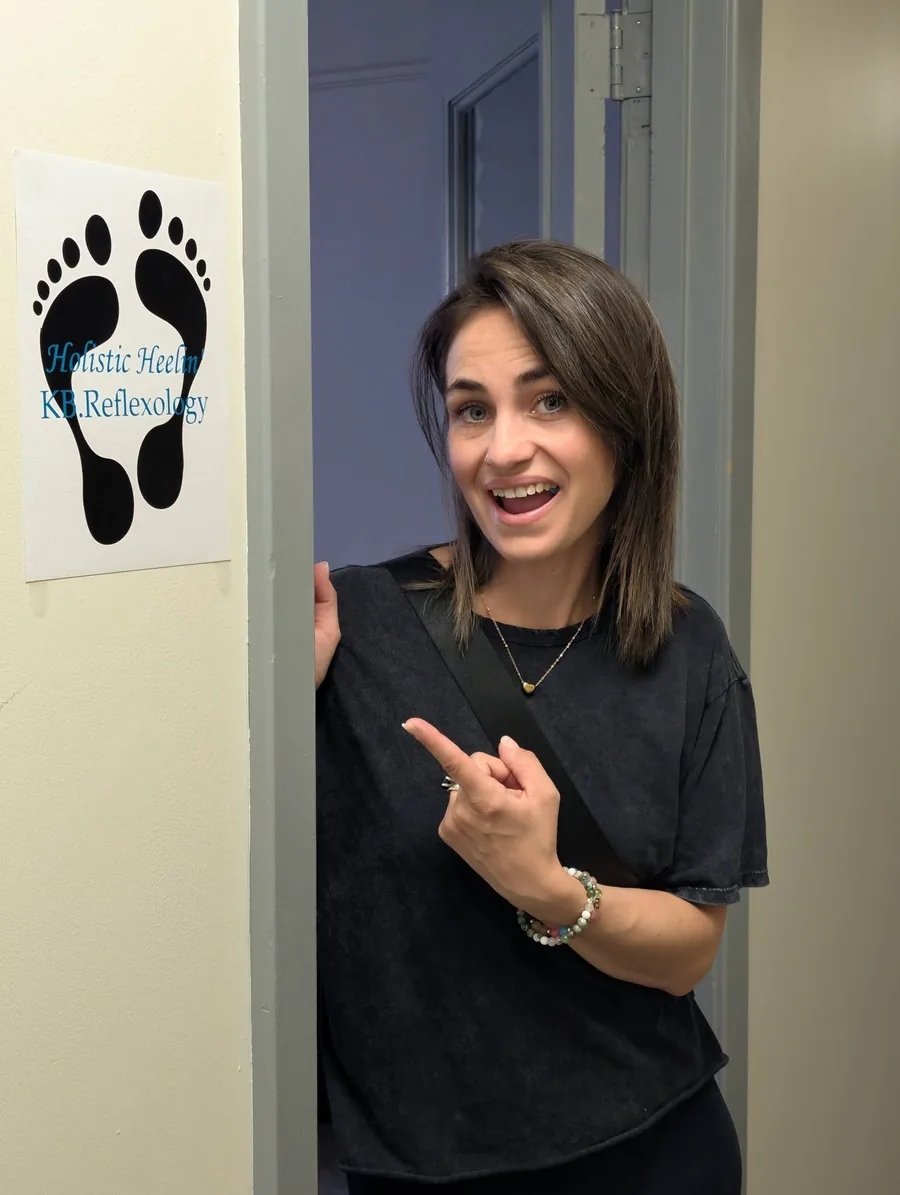 Krista in the doorway of her treatment room, pointing at the sign beside it showing two black footprints and the words Holistic Heelin' KB Reflexology.