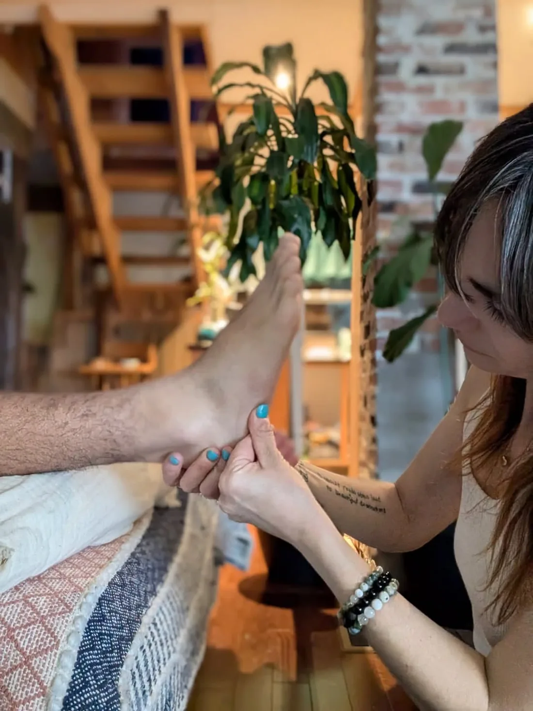 Krista working on a client's foot with both hands, in a warm room with a plant and exposed brick behind her.