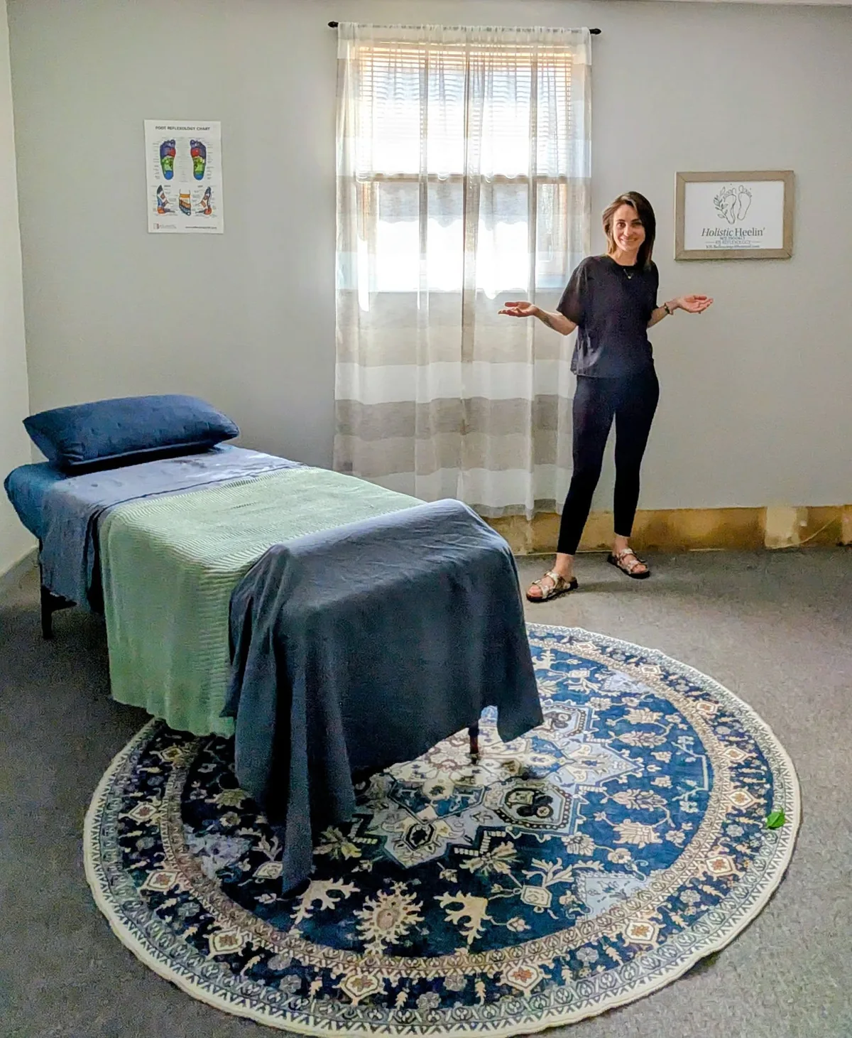 Krista standing beside the treatment table in her room, with a foot reflexology chart on the wall, a curtained window and a round patterned rug.